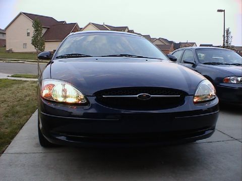 2000 Ford Taurus - A dark blue 2000 Ford Taurus is parked in a driveway, facing the camera with its headlights illuminated.