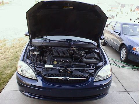 2000 Ford Taurus - Open engine bay of a dark blue Ford Taurus showing the V6 intake manifold, parked in a driveway beside another sedan.