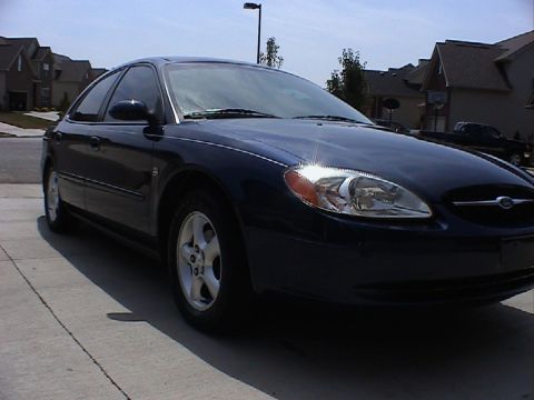 2000 Ford Taurus - A dark blue 2000 Ford Taurus sedan is parked on a concrete driveway in front of houses.