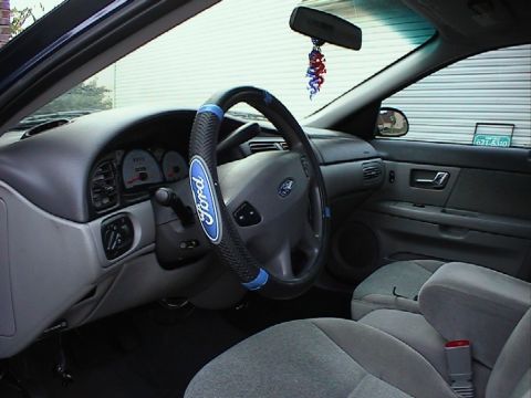 2000 Ford Taurus - Ford sedan cabin showing a Ford-branded padded wheel cover, gray dash and cloth bucket seats, with a red-white-blue hanging ornament.