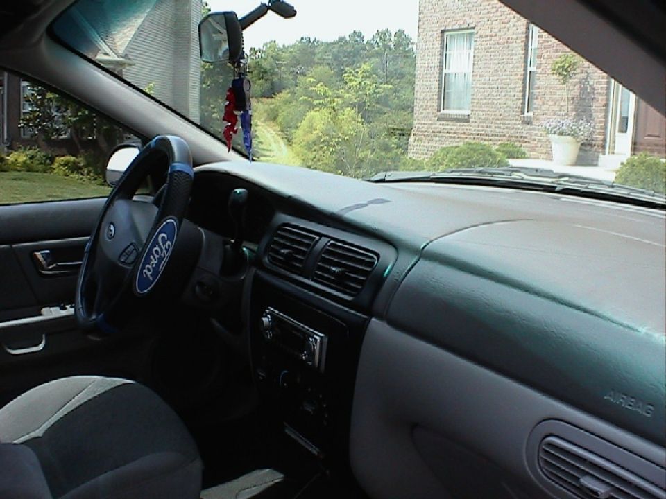 Car interior showing dashboard, steering wheel with Ford logo, and center console. Outside view shows a brick house and trees