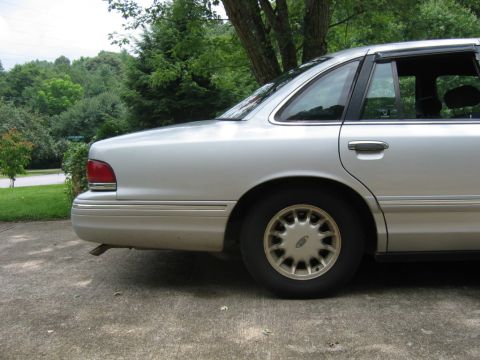 1985 Ford LTD Crown Victoria - Silver sedan parked on a concrete driveway with trees and a hill in the background.