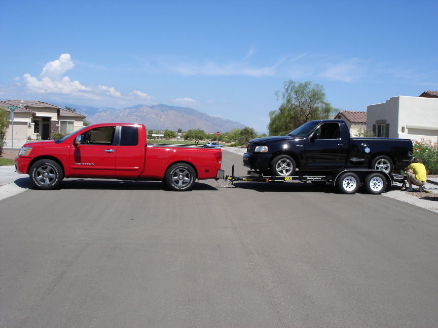 Red Nissan Titan truck towing a black Ford pickup on a flatbed trailer