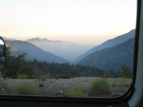 1990 Ford Bronco - Layered forested mountain ridges fade into haze at dusk, framed through an open vehicle window with a side mirror visible.