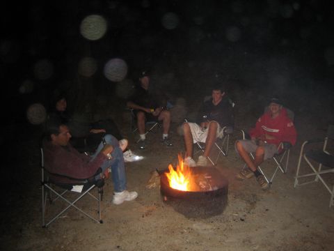 1990 Ford Bronco - Five people relax in folding camp chairs around a glowing fire pit at night, with camera flash dust specks in the dark air.