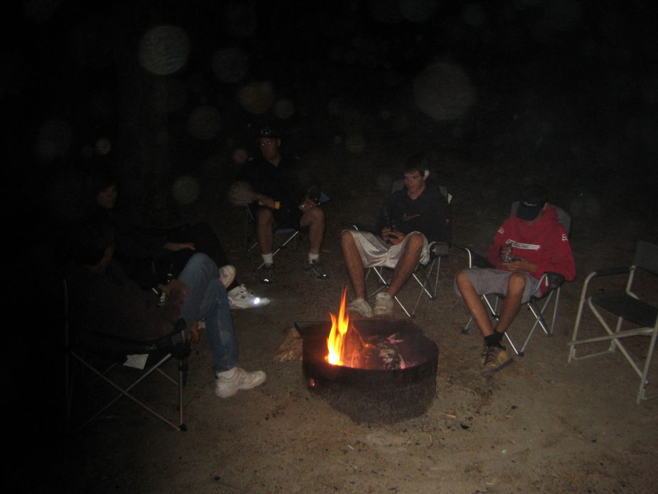 People in camp chairs around a fire pit at night, one in a red jacket