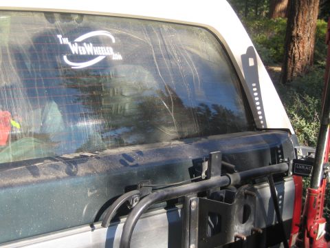1990 Ford Bronco - dusty rear window of an SUV bears a TheWebWheeler.com decal above a tube spare-tire swing-out carrier in a forest.