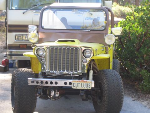 1990 Ford Bronco - bright yellow flat-fender Jeep with a chrome slotted grille and round headlights sits parked beside a motorhome, plate reading CUT LUUS.