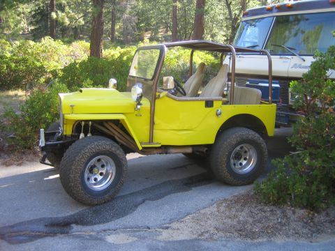 1990 Ford Bronco - lifted bright yellow open-top flat-fender Jeep with chrome wheels and tan seats sits on pavement beside a motorhome among pines.