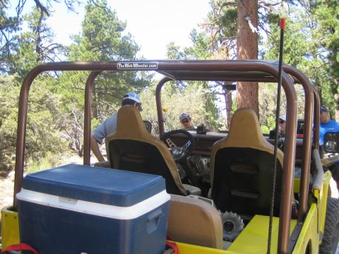 1990 Ford Bronco - View from behind an open Jeep's tan bucket seats and roll cage, a blue cooler strapped at the rear, with people and pines around.
