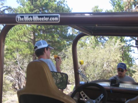 1990 Ford Bronco - man in a white cap sips a drink in the seat of an open Jeep marked TheWebWheeler.com, another man nearby among pine trees.