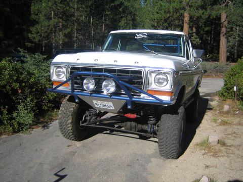 1990 Ford Bronco - lifted silver Ford pickup with FORD lettered grille, tubular bumper, round driving lights and big tires sits parked among pine trees.