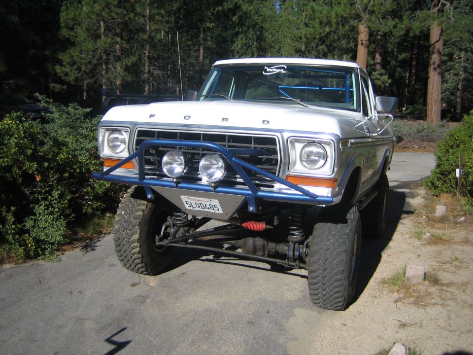 Front of a lifted silver Ford pickup with tube bumper and round lights in pines