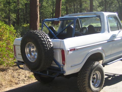 1990 Ford Bronco - lifted white Ford Bronco with an open top, blue interior roll cage, and rear-mounted spare tire sits parked among pines.