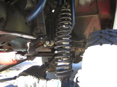 1990 Ford Bronco - close-up underneath a vehicle shows a black coil spring, control arms, steering linkage with a red boot, and a mud tire.