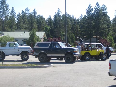 1990 Ford Bronco - lifted black Ford Bronco, a white pickup, and a yellow Jeep gather with a group of people in a tree-lined parking lot.