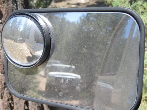 1990 Ford Bronco - dusty vehicle side mirror with a convex blind-spot add-on reflects an off-road vehicle on a trail.