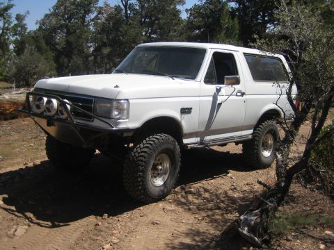 1990 Ford Bronco - white lifted Ford Bronco with a tube front bumper and round driving lights sits on rocky terrain.