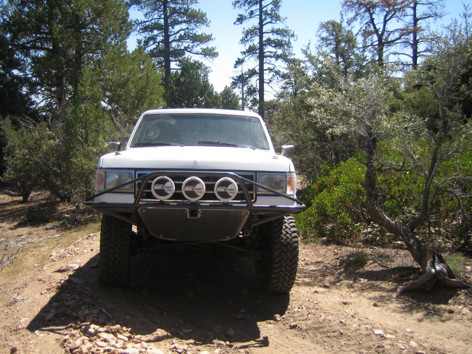 Front view of white lifted truck with tube bumper and three round driving lights on a trail