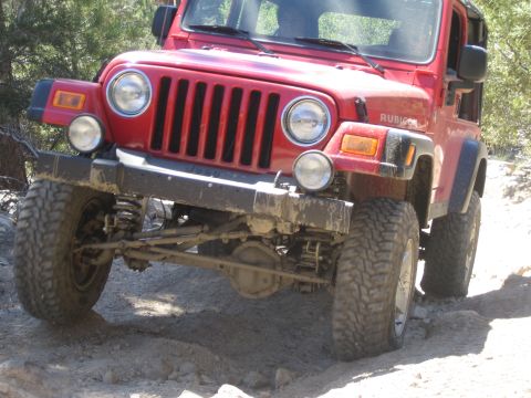 1990 Ford Bronco - Front three-quarter view of a muddy red Jeep Wrangler Rubicon with suspension flexed over a rock ledge.