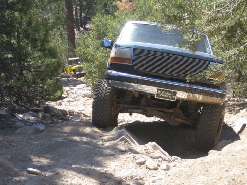 1990 Ford Bronco - dark Ford pickup with a billet grille crawls over a rock ledge as a yellow Jeep waits behind on the trail.