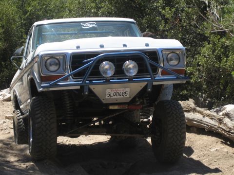1990 Ford Bronco - Front view of a white vintage Ford pickup, FORD lettered on the hood, flexed over rocks on a trail.