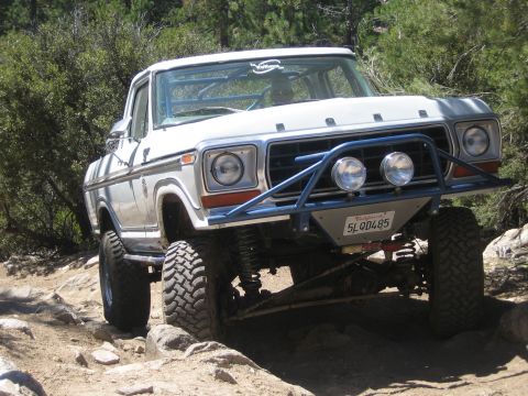 1990 Ford Bronco - white vintage Ford pickup with FORD on the hood and a tube bumper crawls over a rock ledge near brush.