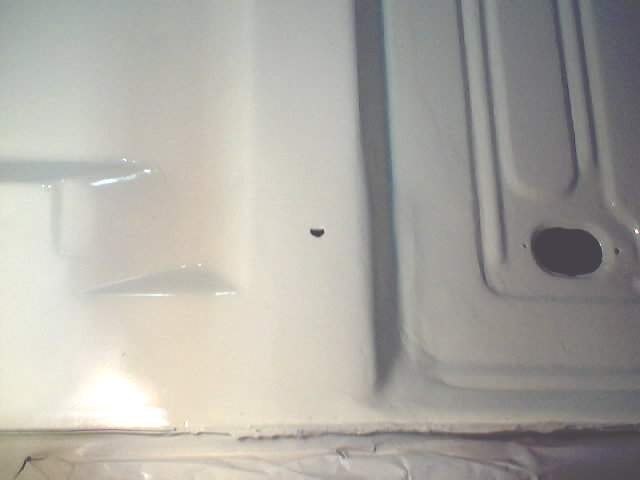 Close-up of a car's floor pan, painted white. Shows some structural ribs and a hole