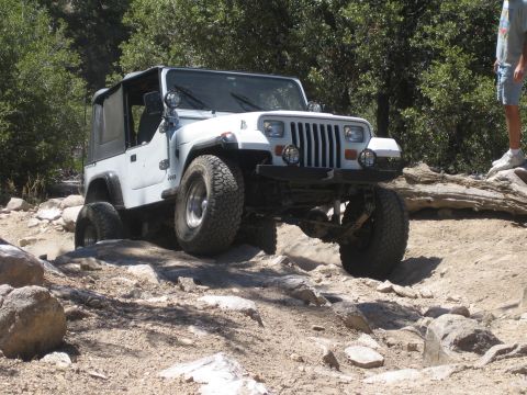1990 Ford Bronco - white square-headlight Jeep Wrangler flexes over pale rocks as a person stands on a log to the side.