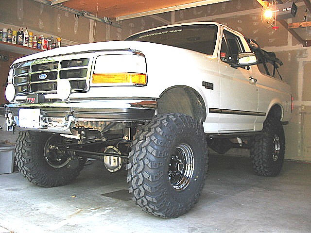 White lifted Ford Bronco on large mud tires parked in a garage, low front 3/4 view.