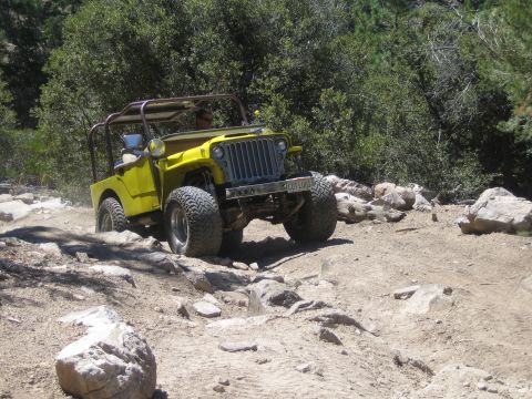 1990 Ford Bronco - yellow flat-fender Willys-style Jeep with an exposed roll bar climbs a rocky dirt trail in the woods.