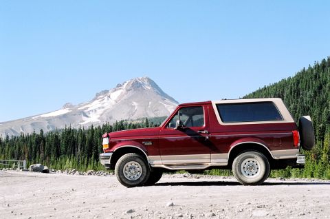 1979 Ford Bronco - Side view of a red Ford Bronco with a tan top, parked in a scenic mountain landscape.