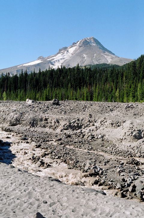 1979 Ford Bronco - A wide shot shows a snow-capped mountain peak above a dense forest and a rocky, dry riverbed.