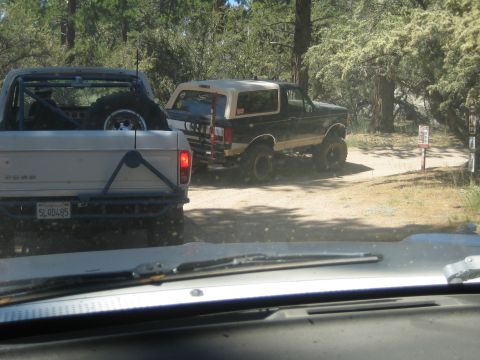 1990 Ford Bronco - Seen through a windshield, a white Ford pickup with spare tire on the tailgate trails a lifted black Bronco on a dusty road.