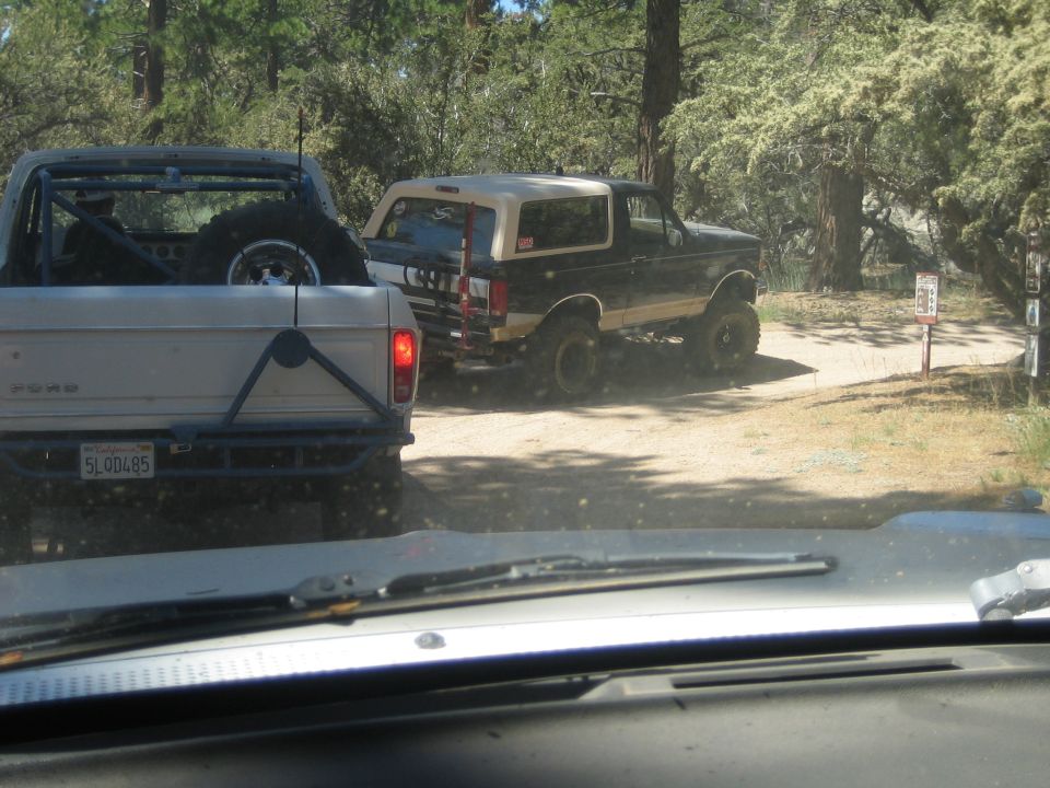View past a Ford pickup tailgate toward a black Bronco on a dirt trail