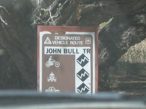 1990 Ford Bronco - brown Forest Service trail sign reads Designated Vehicle Route, John Bull Tr, with motorcycle, ATV, and 4x4 difficulty symbols.