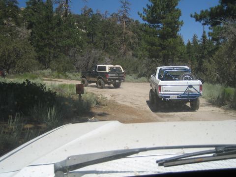 1990 Ford Bronco - Viewed over a dusty hood, a black SUV and a white pickup with roll bar drive a dirt trail through pines and brush.