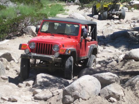 1990 Ford Bronco - red Jeep Wrangler Rubicon climbs large granite boulders on a rocky desert trail, a yellow buggy behind it.