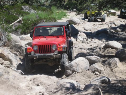 1990 Ford Bronco - red Jeep Wrangler picks its way down a wide boulder field, with a yellow buggy and other rigs in the distance.