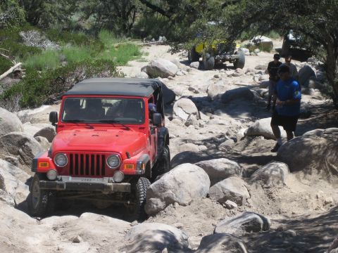 1990 Ford Bronco - red Jeep Wrangler works through boulders while a man in a blue shirt walks alongside spotting, a yellow buggy ahead.