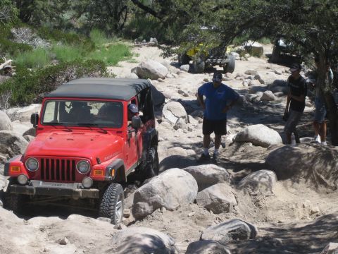 1990 Ford Bronco - red Jeep Wrangler Rubicon sits among boulders as two men in shorts stand on the rocks watching.