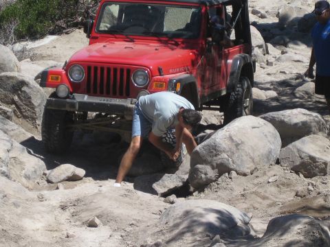 1990 Ford Bronco - man in a gray shirt crouches and inspects the front wheel of a red Jeep Wrangler stopped on a rocky trail.
