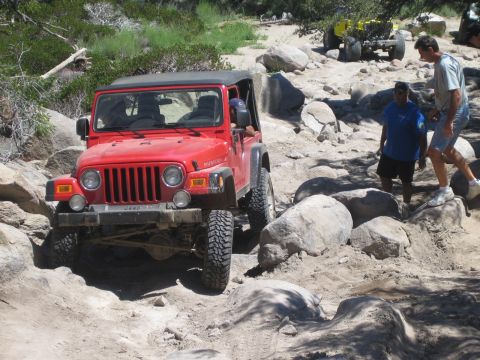 1990 Ford Bronco - red Jeep Wrangler Rubicon eases over boulders while two men stand on the rocks alongside watching the line.