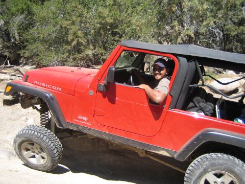 1990 Ford Bronco - smiling man in a cap drives a red soft-top Jeep Wrangler Rubicon Unlimited on a sandy trail.