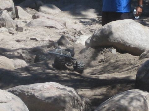 1990 Ford Bronco - miniature silver radio-controlled monster truck on oversized tires crawls between large rocks as a person stands behind.