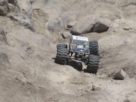 1990 Ford Bronco - small silver radio-controlled monster truck on four large tires climbs a rutted dirt slope.