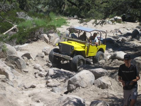 1990 Ford Bronco - yellow vintage flat-fender Jeep with a white hardtop crawls over boulders as a man in a black shirt walks past.