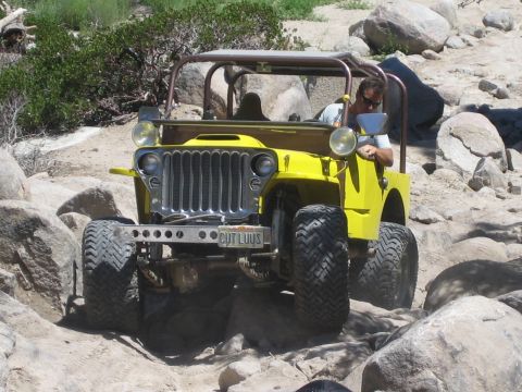1990 Ford Bronco - yellow flat-fender Jeep with an exposed roll cage drops over boulders, its front plate reading CUT LUUS.