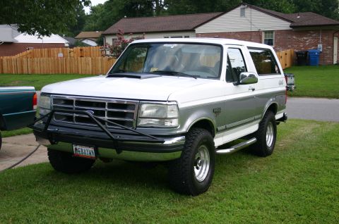 1994 Ford Bronco - White-over-silver two-tone Ford Bronco parked on a lawn in a three-quarter front view, with houses and a wood fence behind it. It has a bla…