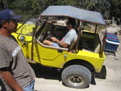 1990 Ford Bronco - man drives a yellow flat-fender Jeep with a metal roof while another man in a cap stands beside it on a dusty trail.